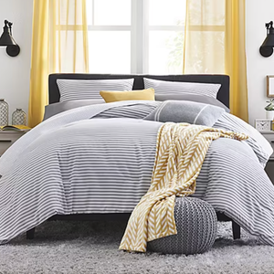 A cozy king-sized reversible comforter set with gray and white stripes, complemented by striped pillows and a touch of yellow from a blanket and pillow. The bed is framed with yellow curtains and rests on a textured gray rug.