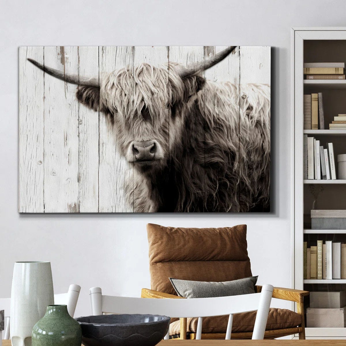 A black and white canvas features a Highland cow with long hair and horns, set against a white wooden background, hanging above a cozy chair and bookshelf.