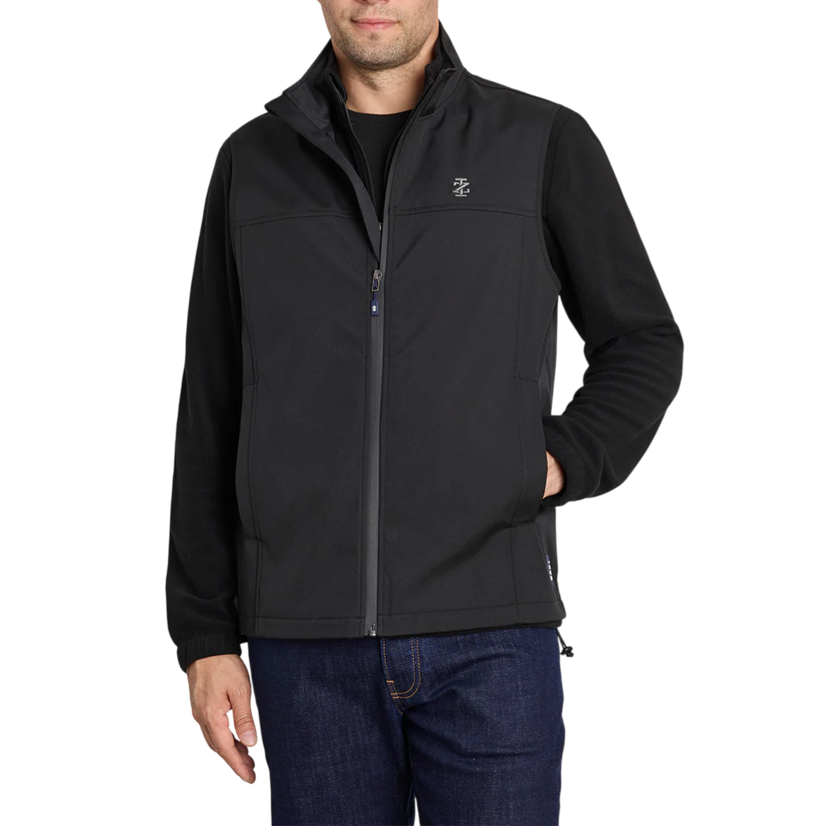 A man is wearing a black softshell jacket with a front zipper and a small logo on the chest, paired with dark jeans.