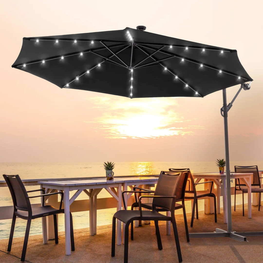 A large black patio umbrella with built-in LED lights is set up over an outdoor dining table and chairs, facing a sunset ocean view.