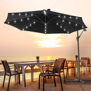 A large black patio umbrella with built-in LED lights is set up over an outdoor dining table and chairs, facing a sunset ocean view.