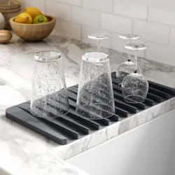 A black silicone dish drying mat holds two speckled drinking glasses and two upside-down wine glasses, set on a marble kitchen countertop. A bowl of fruit is nearby.