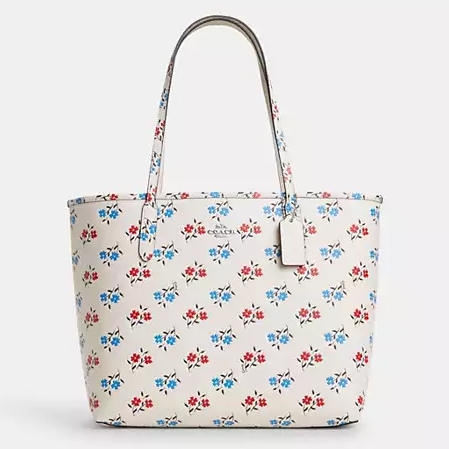 White tote bag featuring a blue and red floral print with two leather handles and a small Coach logo tag attached to the side.