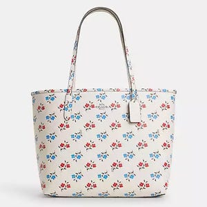 White tote bag featuring a blue and red floral print with two leather handles and a small Coach logo tag attached to the side.
