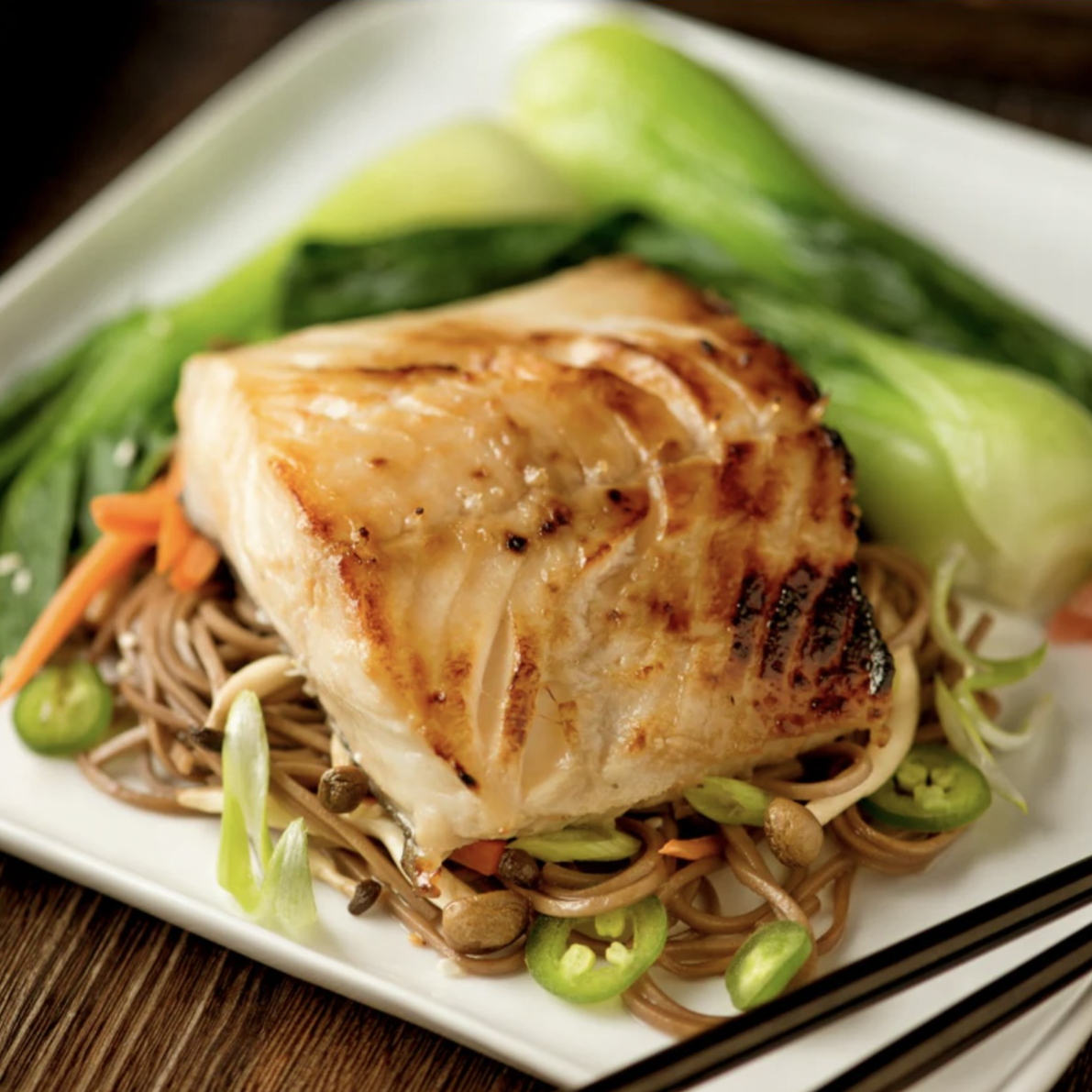 A dish with grilled fish on a bed of soba noodles, garnished with sliced peppers, carrots, and green onions, served alongside bok choy and a side of pickled ginger.