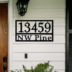 House number sign with the address \“13459 NW Pine\“ displayed on a white exterior wall, featuring black metal design.
