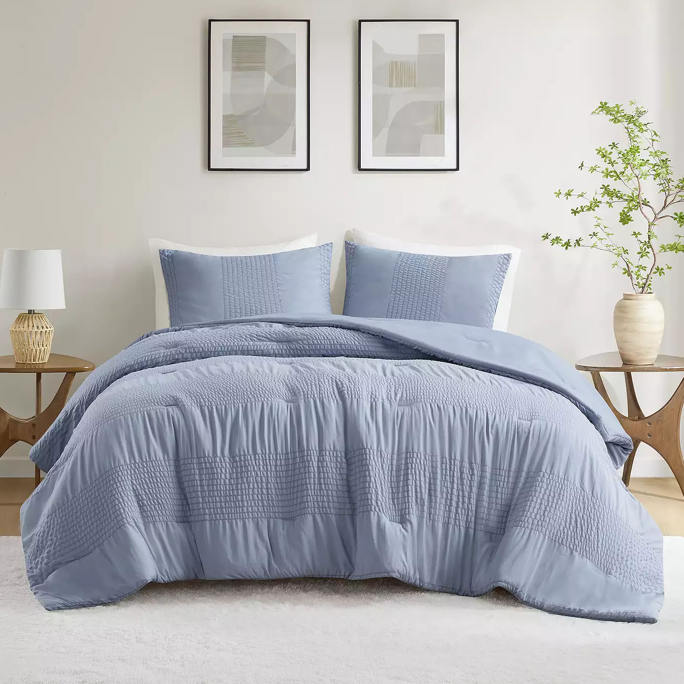 A cozy textured comforter set in a soft blue hue featuring a quilted pattern, accompanied by matching pillow shams. The bedding is styled in a minimalist bedroom with neutral accents and framed abstract art on the wall.