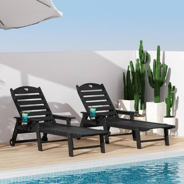 Two black outdoor lounge chairs with heart cutouts on the backrests are placed on a wooden deck beside a swimming pool. Tall cacti in white planters are situated in the background, and blue drinks rest on the chairs' armrests.