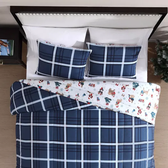A bed is made up with a blue plaid comforter and matching pillow shams. The bedding is turned to reveal a fun, holiday-themed bedsheet with snowmen and reindeer.