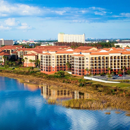 A large hotel complex with multiple floors and distinctive red and beige architecture is located by a calm waterfront, set against a backdrop of urban buildings and a clear sky.