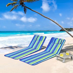 Two colorful, striped, foldable beach lounge chairs are set on the sand beside a low, small table with two glasses filled with a yellow beverage, all under a palm tree by the sea.