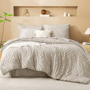 A beige bedding set with a textured chevron pattern, including pillows and a duvet cover, styled in a minimalist bedroom with neutral decor, green plant, books, and decorative sculpture.