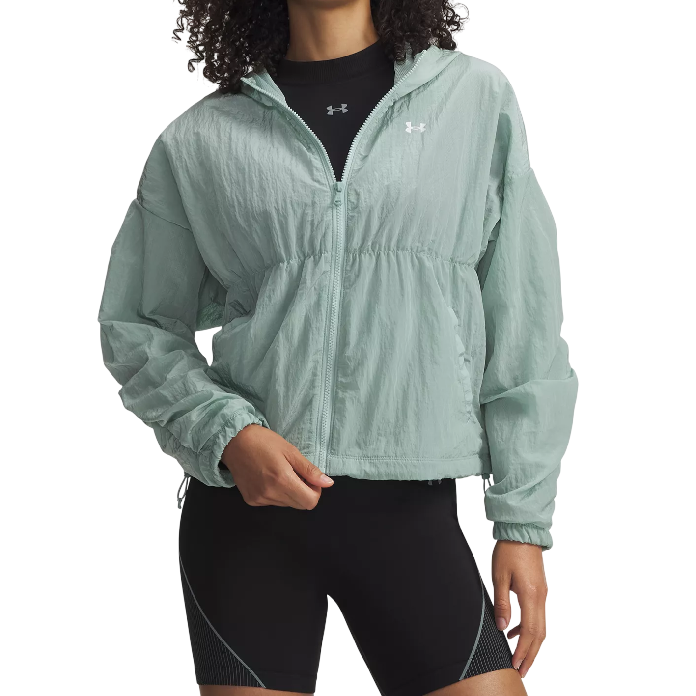 A person wearing a light green Under Armour windbreaker jacket with a zip-up front, layered over a black Under Armour top and paired with black sports shorts.
