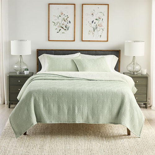 A neatly made bed features a light green quilt and pillow shams with a geometric pattern, flanked by two bedside tables with white lamps and decorated with framed floral artwork above.