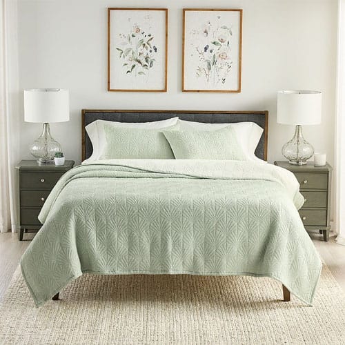 A neatly made bed features a light green quilt and pillow shams with a geometric pattern, flanked by two bedside tables with white lamps and decorated with framed floral artwork above.