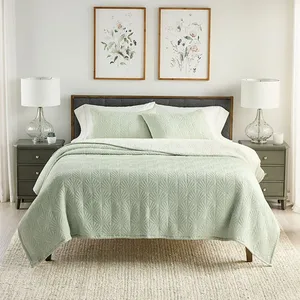 A neatly made bed features a light green quilt and pillow shams with a geometric pattern, flanked by two bedside tables with white lamps and decorated with framed floral artwork above.
