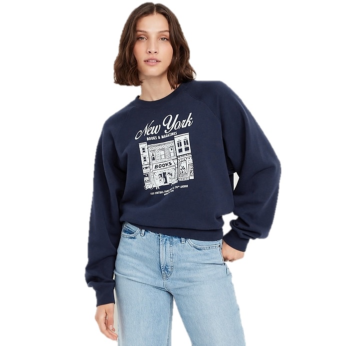 A woman is wearing a navy blue sweatshirt featuring a \“New York Books & Magazines\“ graphic, paired with light blue jeans.