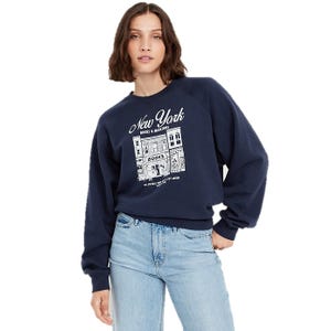 A woman is wearing a navy blue sweatshirt featuring a \“New York Books & Magazines\“ graphic, paired with light blue jeans.