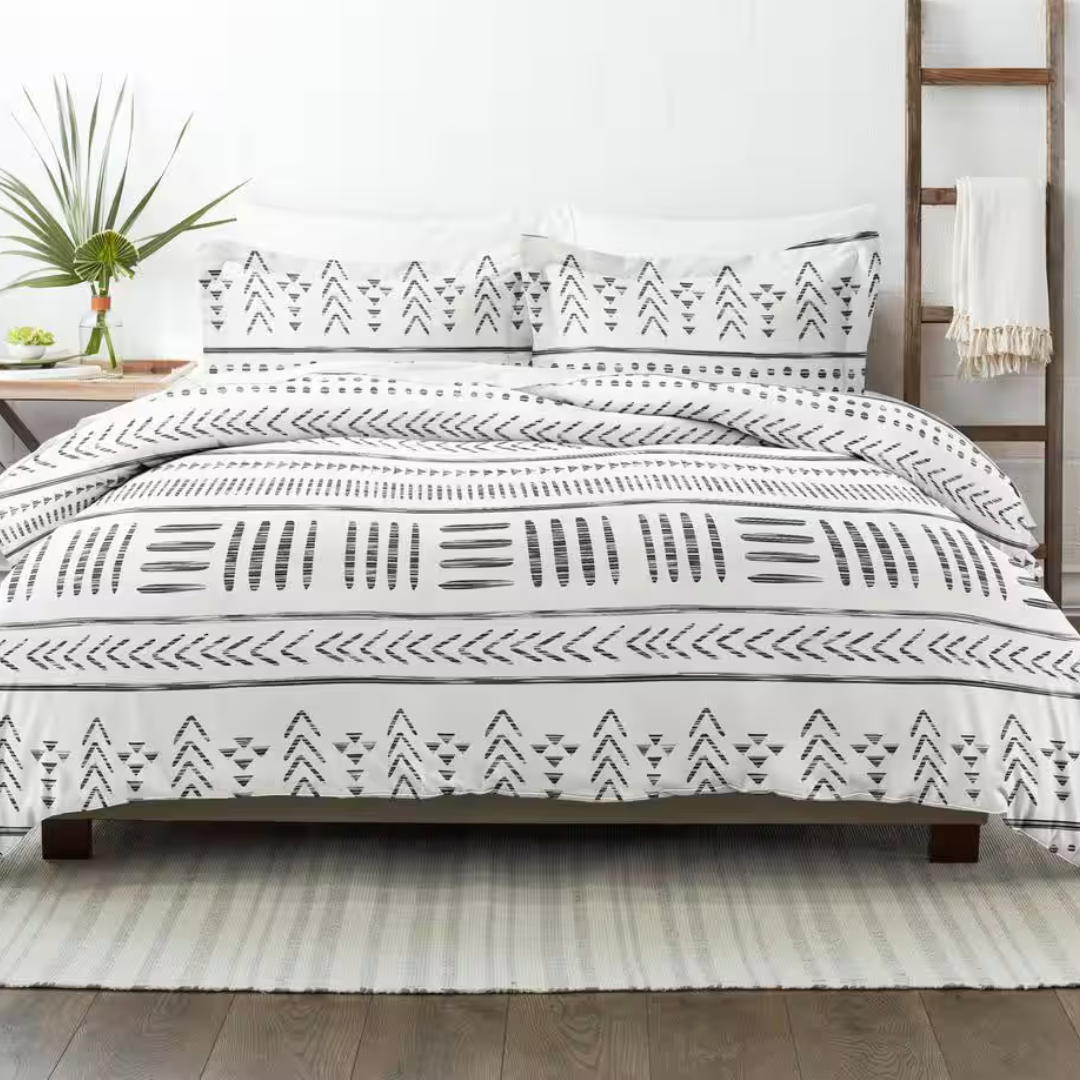 White bedding set with black geometric patterns featuring triangles and lines, placed on a bed. The room includes a plant, a wooden ladder with a throw, and a neutral area rug on a wooden floor.