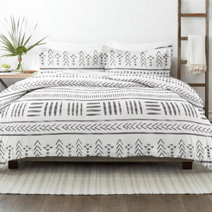 White bedding set with black geometric patterns featuring triangles and lines, placed on a bed. The room includes a plant, a wooden ladder with a throw, and a neutral area rug on a wooden floor.