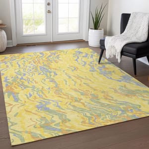 A colorful abstract rug with yellow and multicolored patterns is placed on a wooden floor in a bright room with a chair draped in a white blanket and a potted plant nearby.