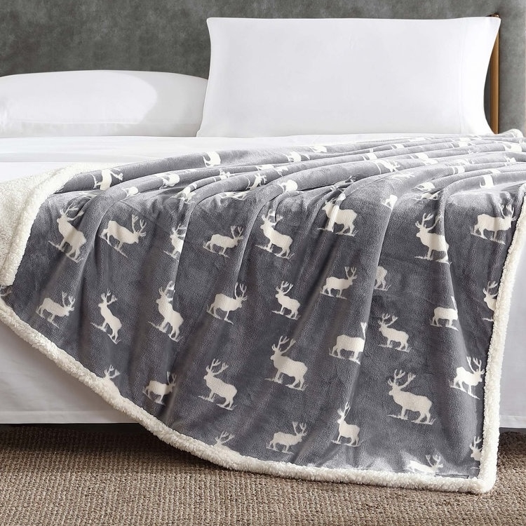 A gray fleece blanket with a white deer pattern is draped over a bed with white pillows.