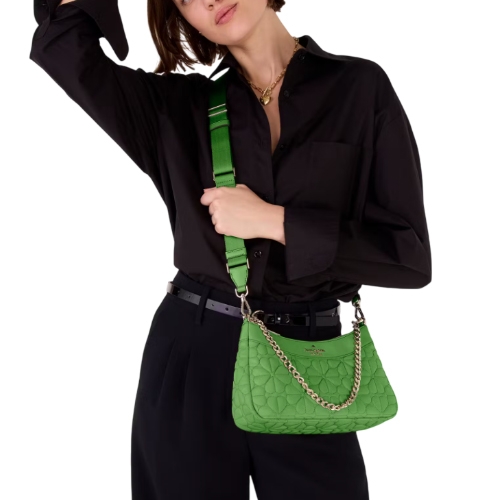 A green Kate Spade quilted crossbody bag features a chain and adjustable strap for versatility, worn by a model in a black outfit, adding contrast and elegance to the ensemble.