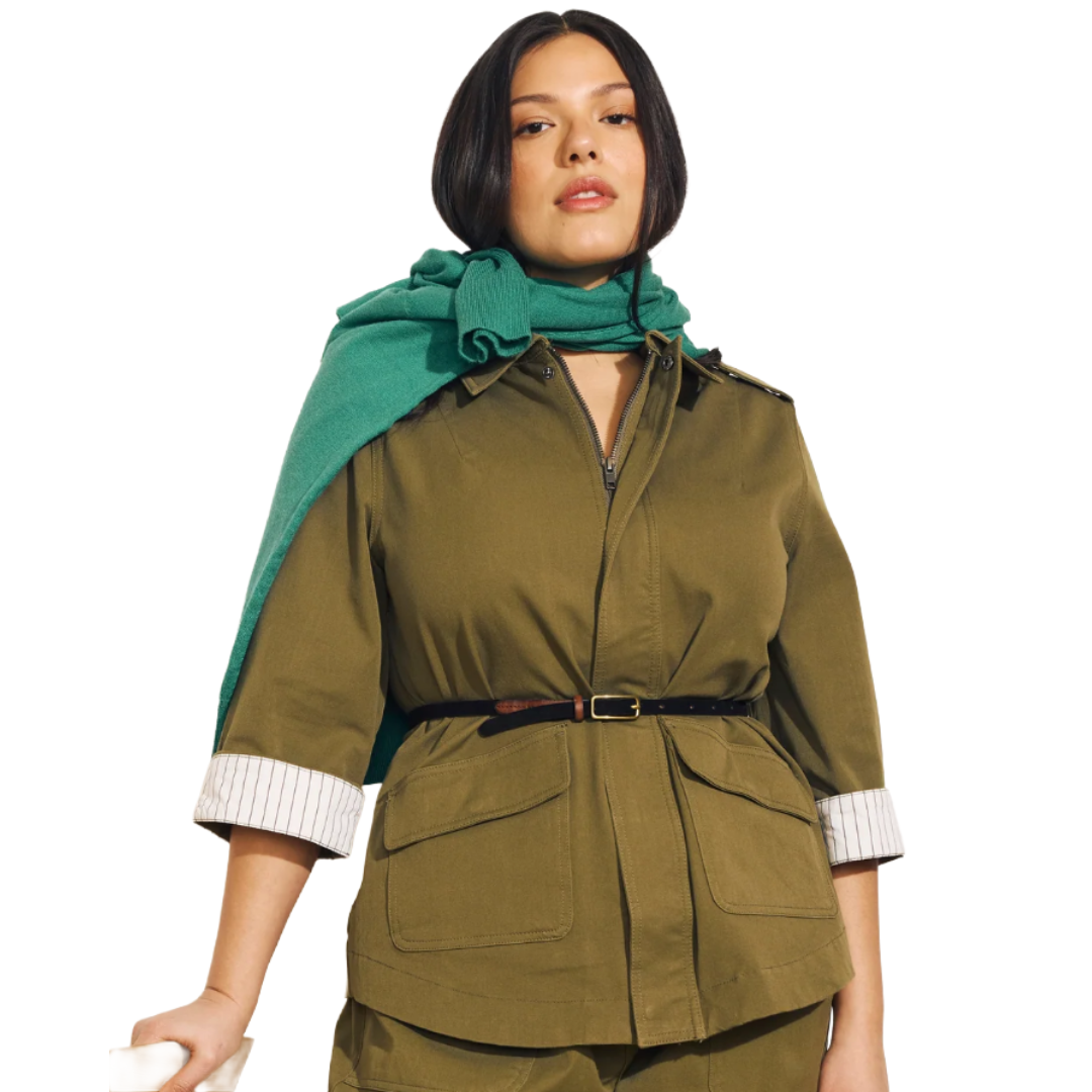 A person is wearing an olive green utility jacket with a belted waist and large front pockets, paired with matching pants. A turquoise sweater is draped over their shoulders.