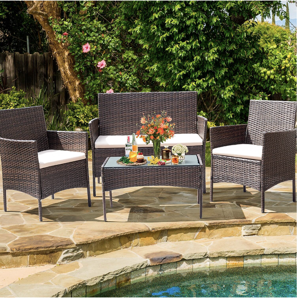 A wicker patio furniture set with cream cushions includes a bench, two chairs, and a glass-top coffee table, placed near a pool with a floral arrangement and drinks on the table.