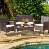 A wicker patio furniture set with cream cushions includes a bench, two chairs, and a glass-top coffee table, placed near a pool with a floral arrangement and drinks on the table.