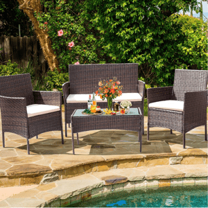 A wicker patio furniture set with cream cushions includes a bench, two chairs, and a glass-top coffee table, placed near a pool with a floral arrangement and drinks on the table.
