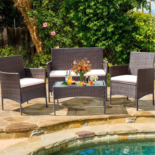 A wicker patio furniture set with cream cushions includes a bench, two chairs, and a glass-top coffee table, placed near a pool with a floral arrangement and drinks on the table.