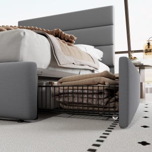 A modern bed with a grey upholstered headboard and footboard features an under-bed storage basket holding folded blankets, in a room with a light carpet and a decorative lamp on a side table.
