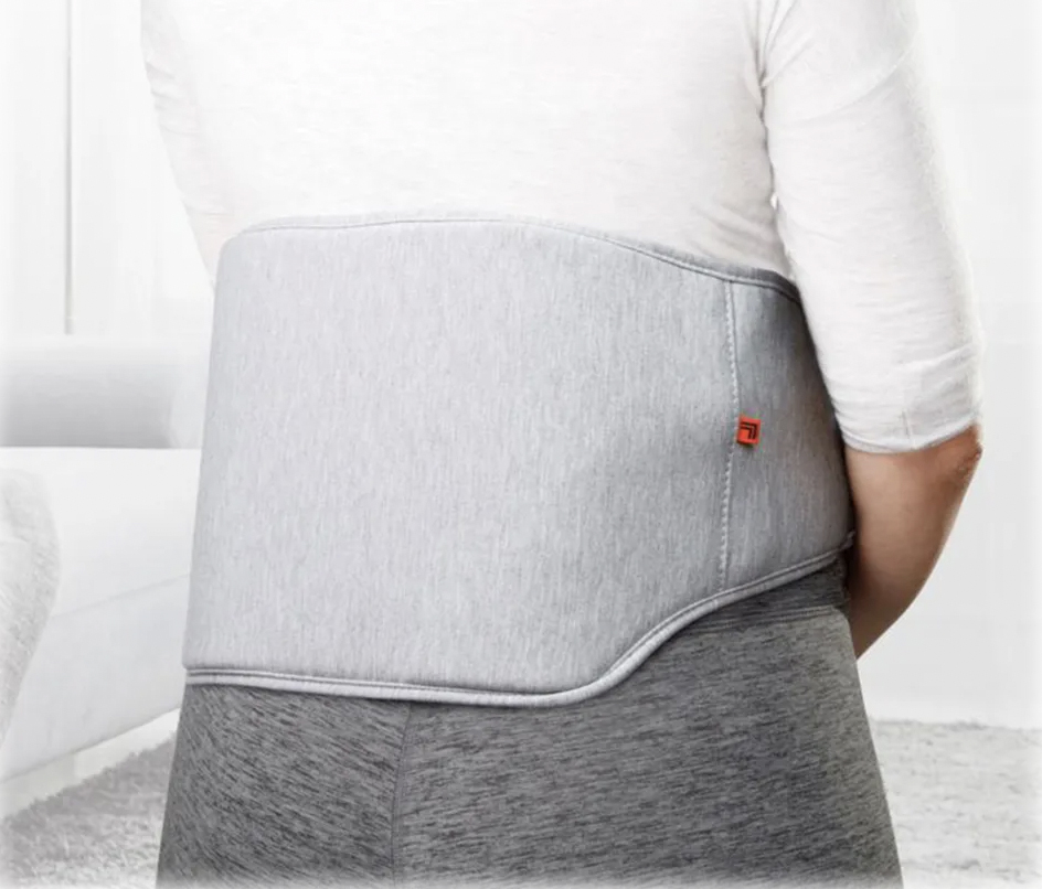 A person is wearing a gray, wrap-around heated back massager designed to provide warmth and massage the lower back area. It features a subtle two-tone design and a small red logo.