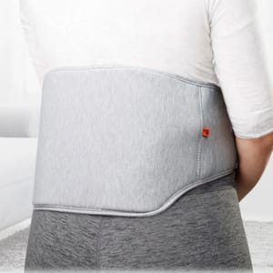A person is wearing a gray, wrap-around heated back massager designed to provide warmth and massage the lower back area. It features a subtle two-tone design and a small red logo.