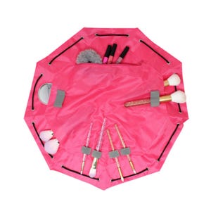 A pink toiletry cinch bag with multiple compartments holds various makeup items, including brushes, lip products, and a compact mirror. The bag features a drawstring closure for easy packing.