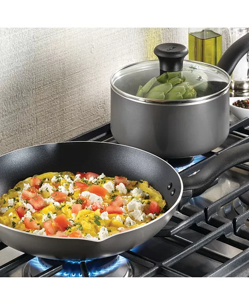 A nonstick fry pan with a vibrant omelette topped with tomatoes and feta, alongside a saucepan with a glass lid containing vegetables, both on a gas stovetop.