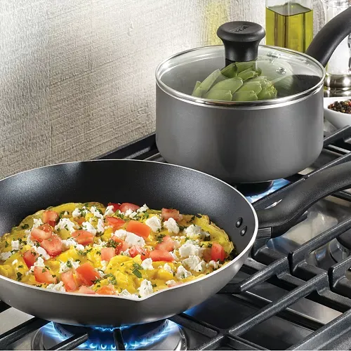 A nonstick fry pan with a vibrant omelette topped with tomatoes and feta, alongside a saucepan with a glass lid containing vegetables, both on a gas stovetop.