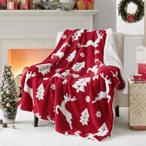 A red and white holiday-themed throw blanket with reindeer and tree designs is draped over a chair, set in a festive living room scene with a decorated Christmas tree, presents, and wreath.