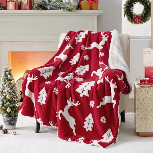 A red and white holiday-themed throw blanket with reindeer and tree designs is draped over a chair, set in a festive living room scene with a decorated Christmas tree, presents, and wreath.