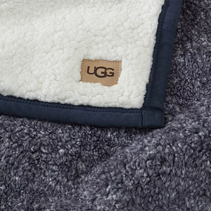 A soft, plush UGG blanket featuring a fluffy white sherpa texture on one side and a dark grey, curly fleece on the other, with a small UGG tag sewn into the material.