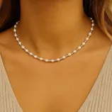 A woman wears a delicate pearl necklace with evenly spaced pearls around her neck. She is dressed in a beige, textured top.
