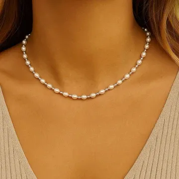 A woman wears a delicate pearl necklace with evenly spaced pearls around her neck. She is dressed in a beige, textured top.