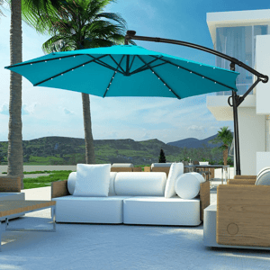 A modern outdoor lounge area features a white sectional sofa with cushions and a wooden coffee table, shaded by a large teal cantilever umbrella. Palm trees and a scenic landscape complete the setting.