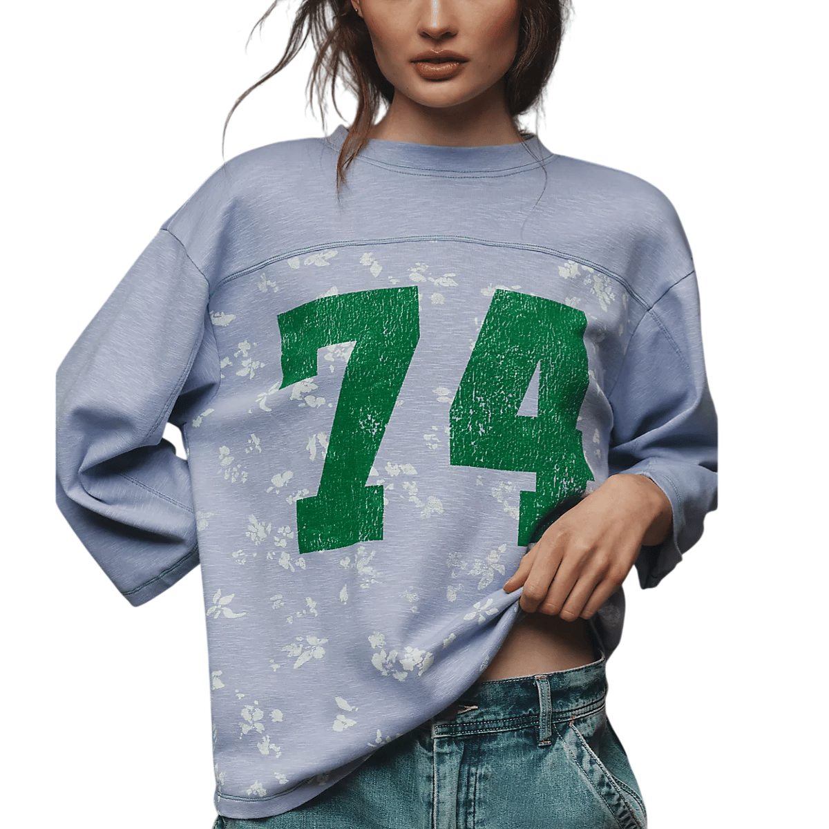 A person is wearing a light blue sweatshirt with the number \“74\“ in large green print, featuring subtle floral patterns. It is paired with blue denim jeans.