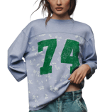 A person is wearing a light blue sweatshirt with the number \“74\“ in large green print, featuring subtle floral patterns. It is paired with blue denim jeans.