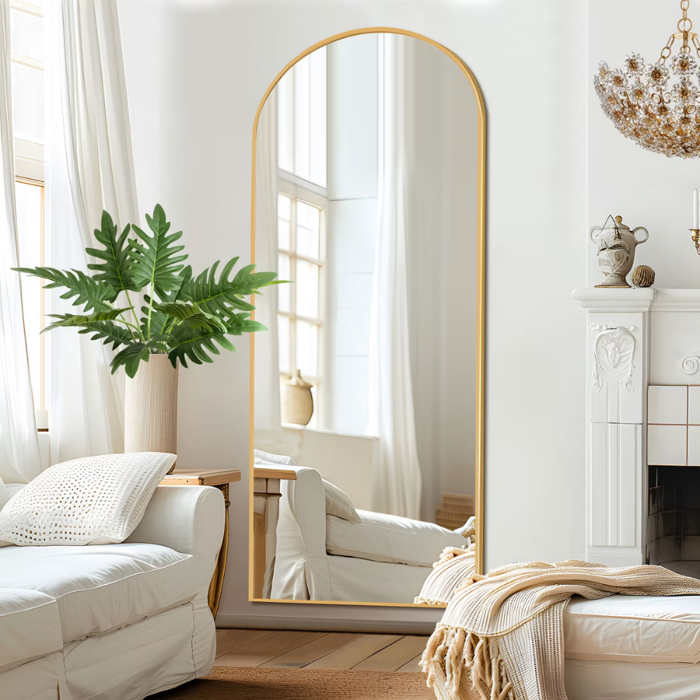 A large, arched mirror with a gold frame is placed in a room featuring a white sofa, a decorative fireplace, a potted plant, a chandelier, and light curtains.