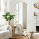 A large, arched mirror with a gold frame is placed in a room featuring a white sofa, a decorative fireplace, a potted plant, a chandelier, and light curtains.