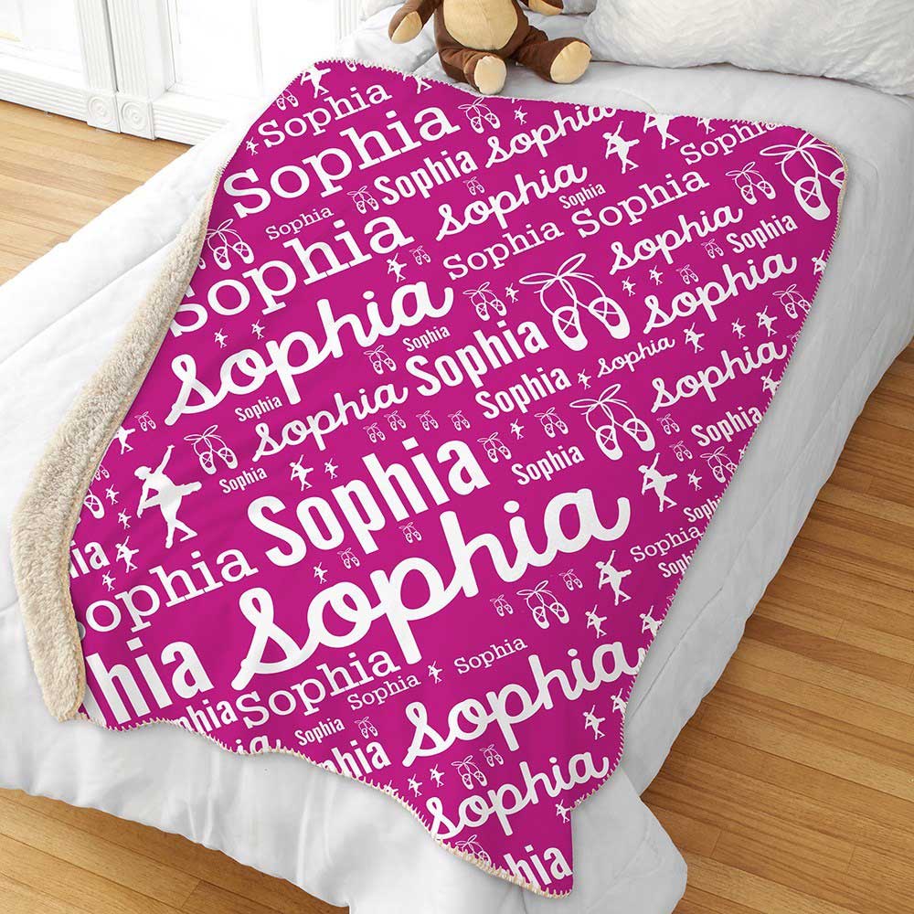 A pink blanket with the name \“Sophia\“ in various fonts and sizes, featuring ballet shoes, butterflies, and dancer silhouettes, is draped over a bed. A stuffed bear sits near the top corner.