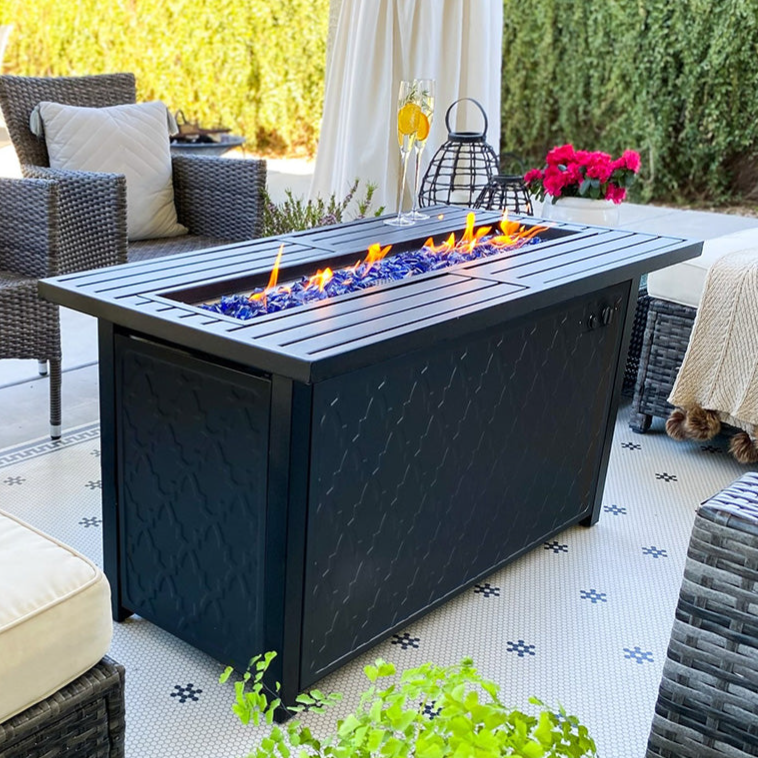 A stylish outdoor patio set with wicker chairs, a cushioned seat, and a sleek rectangular fire pit table featuring blue glass stones. Small potted plants and decorative items add to the inviting atmosphere.
